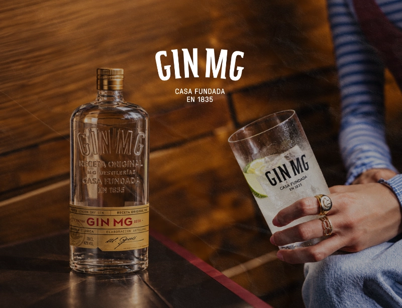 Botella de Gin MG y gin tonic con lima en la mano de una persona sobre una mesa de madera, proyecto de paid media de KLOROFiLA